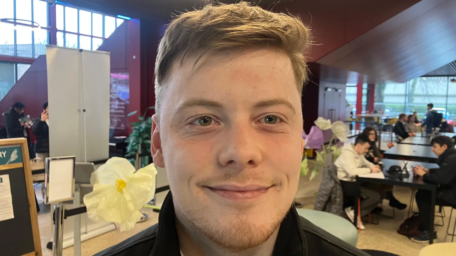 A close-up image of Ben Friel. He is a young man with short, light coloured hair and a beard. He is smiling at the camera. He is standing in front of some fake white and purple flowers and behind him are some students sat at tables.