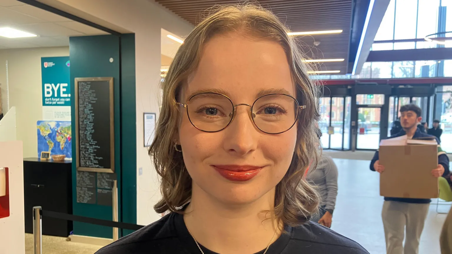 Lucy O'Rawe is a young woman with short, light-coloure hair and she is wearing a pair of circular glasses. She is smiling at the camera. She is also wearing a blue jumper and a thin chain necklace. She is stood in front of The Pantry, with students stood beside her bringing in large cardboard boxes.