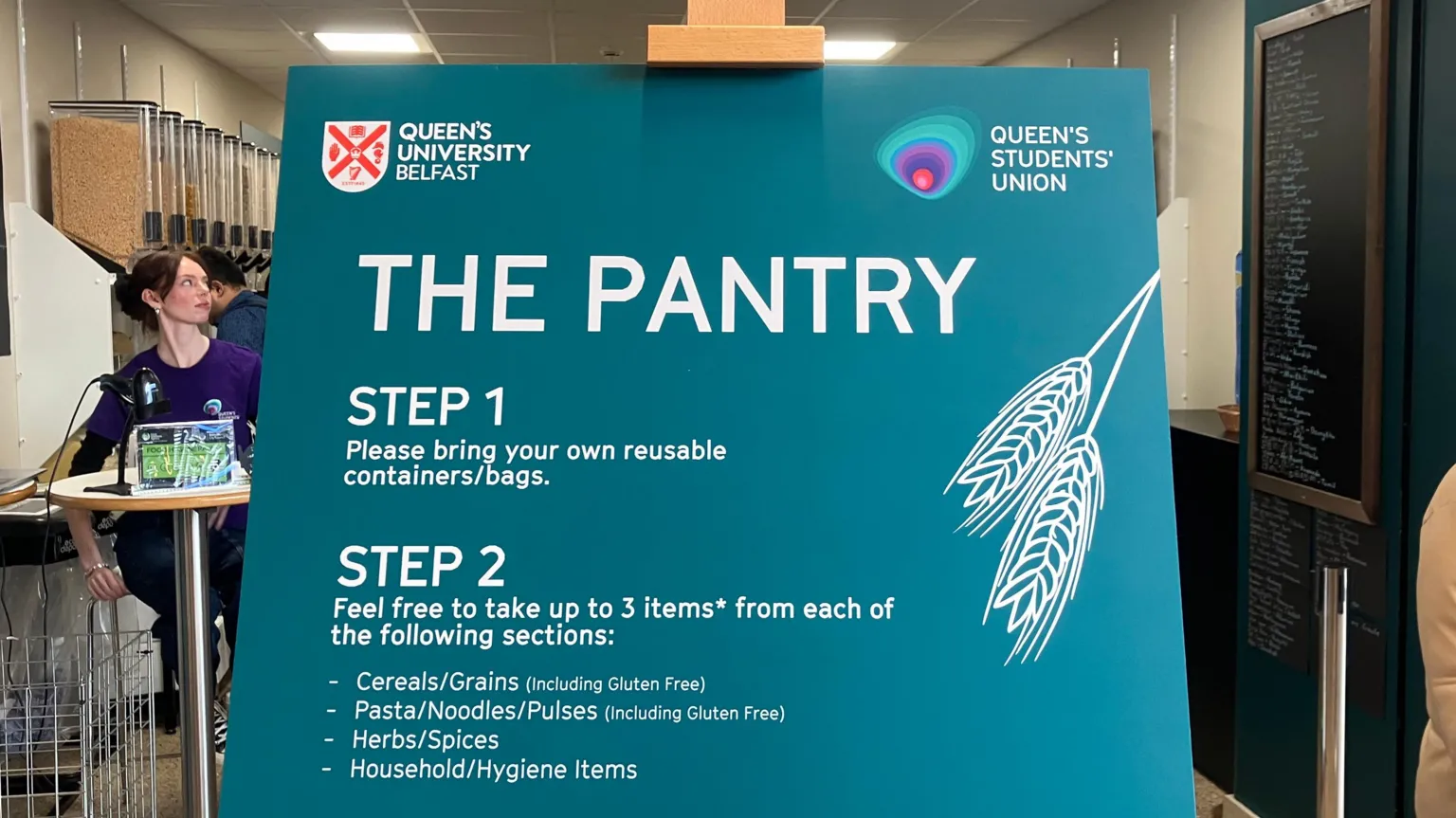A large board for The Pantry is placed on a wooden easel. It shows the steps for using the facility, such as bringing your own reusable container and what items students can avail of.