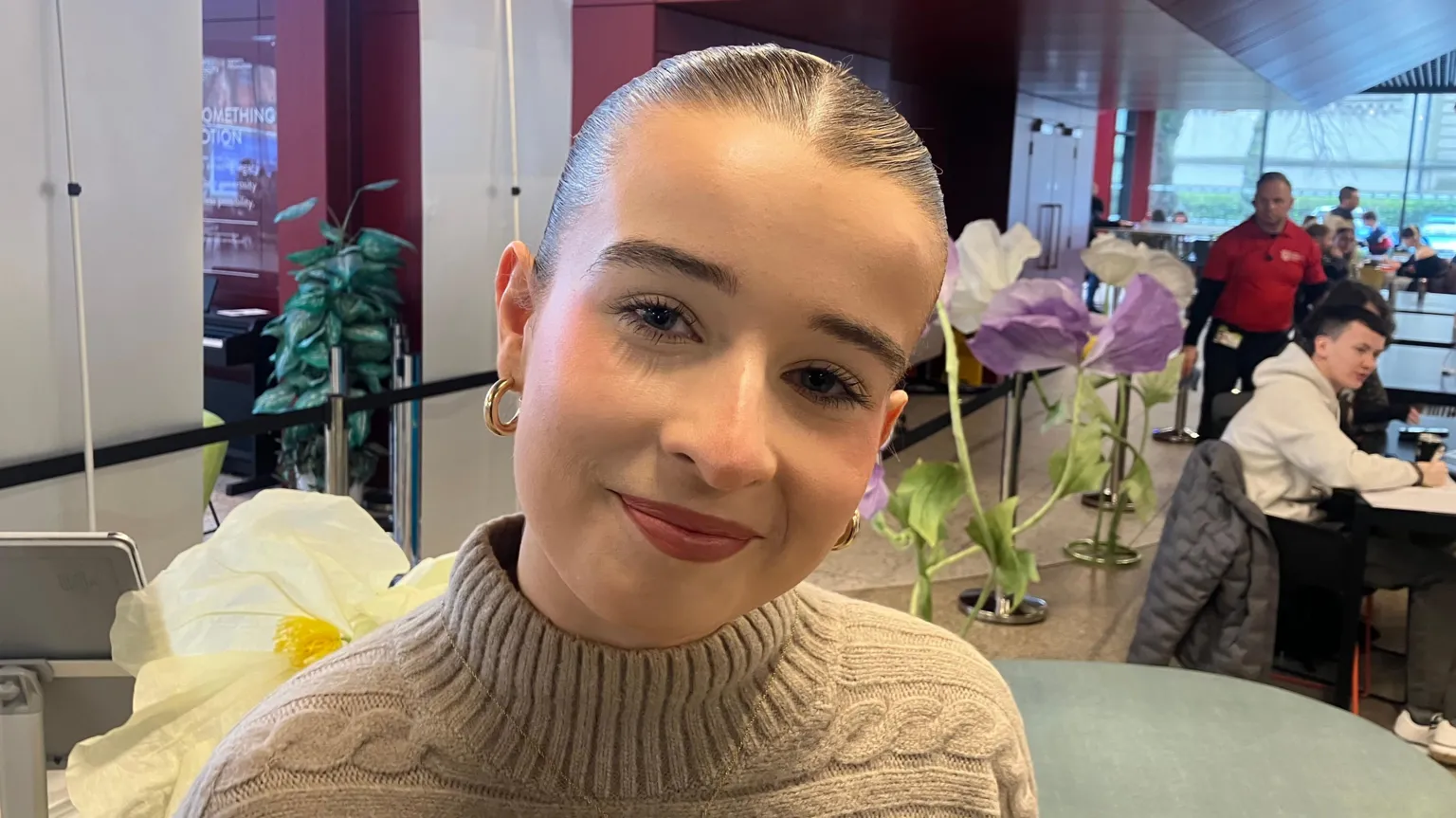 Holly Dougan is a young woman with slicked back, dark hair. She is wearing a beige jumper with knitted patterns on it and a pair of gold hoop earrings. She is standing in the middle of the Students' Union, while others sit at desks in the distance. Holly is smiling at the camera.