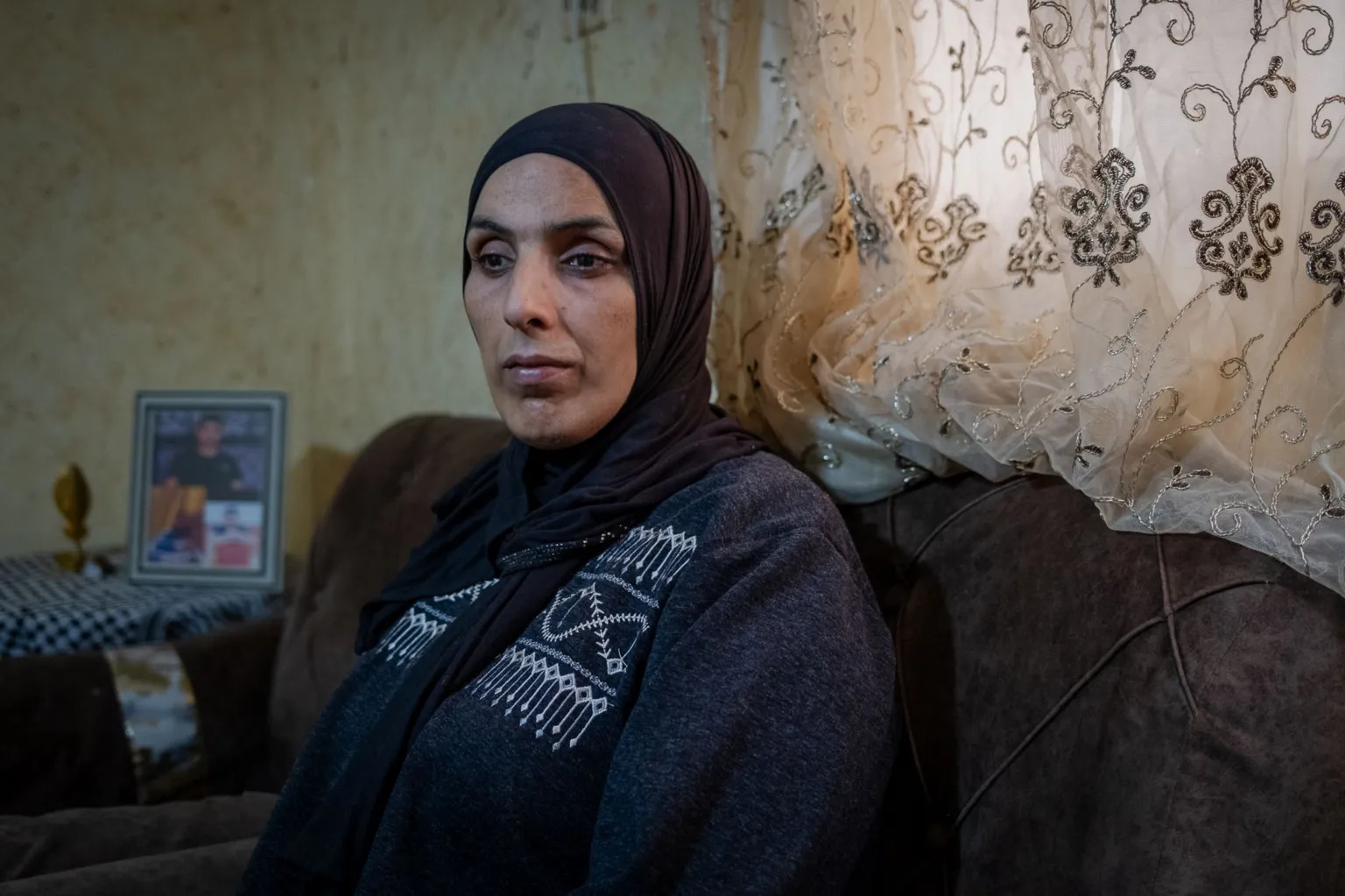 A close up of Jad's mother looking forlorn, sitting in the family living room in al-Far'a refugee camp, in the occupied West Bank. Behind her on the table is a framed picture of Jad.
