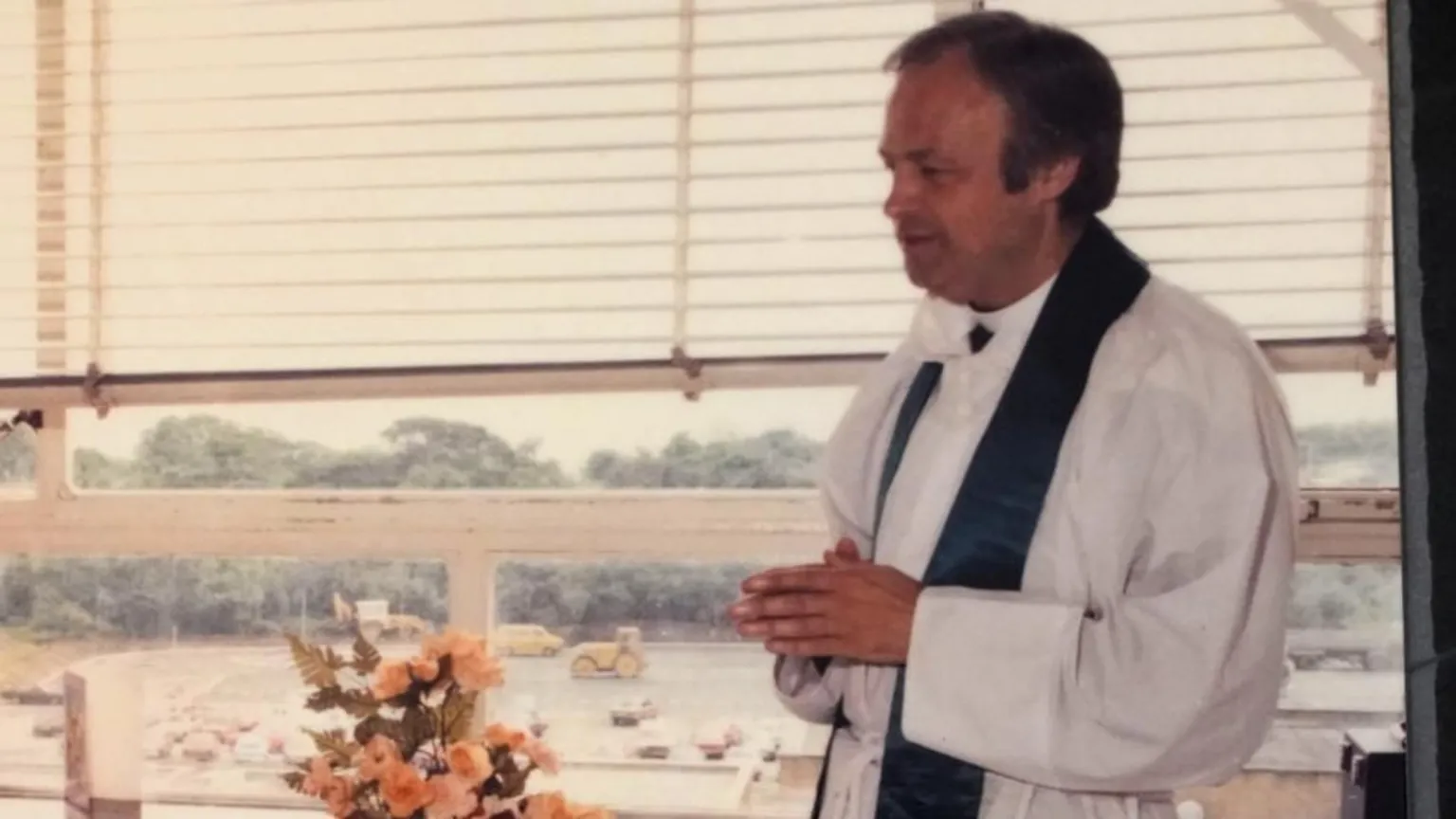 D J Photography A younger Anthony Pierce stands clasping his hands with his back to a window. He is side of frame and is wearing white robes with a dark silk stole around his neck.