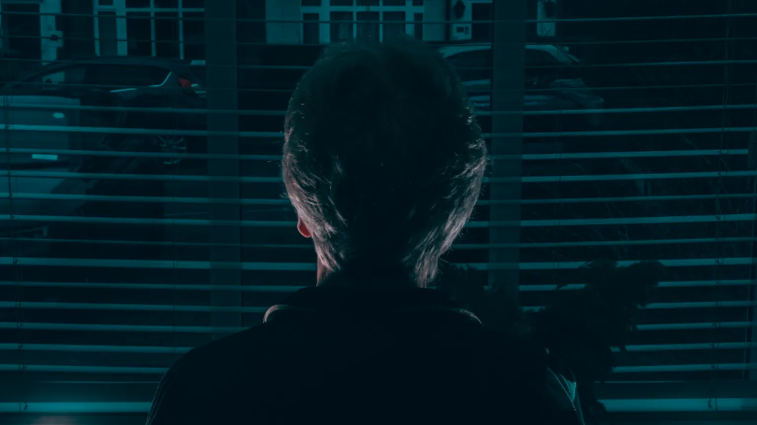 A woman stands looking out of a window. She has short hair and her back to the camera, to protect her identity.