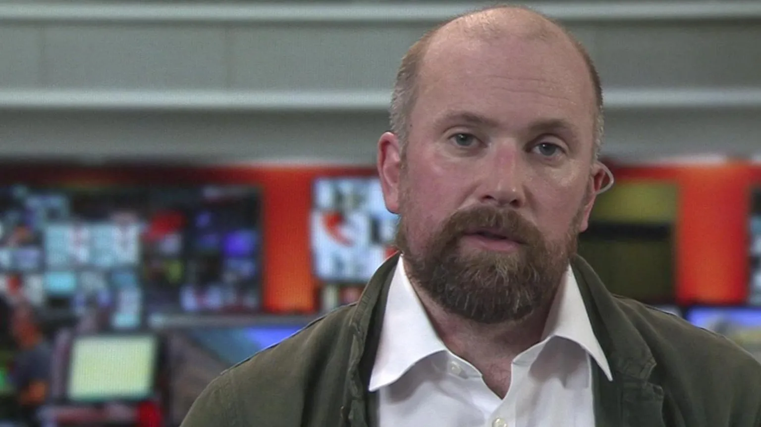 A head and shoulders image of David Shipley. He is wearing a green jacket over a white shirt. He has facial hair and is looking directly into the camera. He appears to be in a television studio, as lots of screens are in the background.