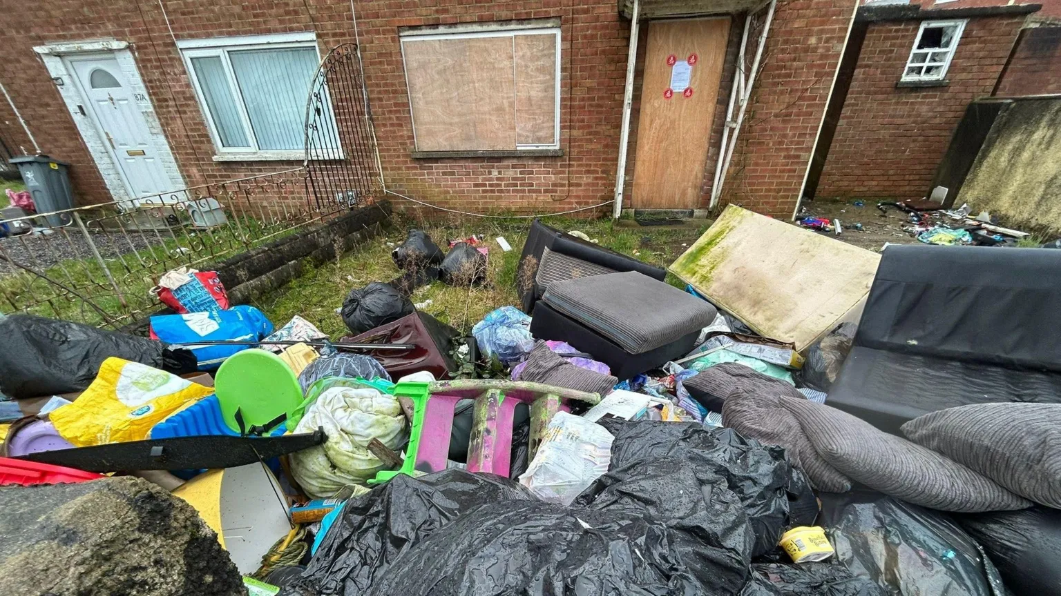 Local Democracy Reporting Service A large pile of filled black bin bags, carrier bags, a sofa and a bed frame and mattress are among the items left in the garden. The front window and door are boarded up.