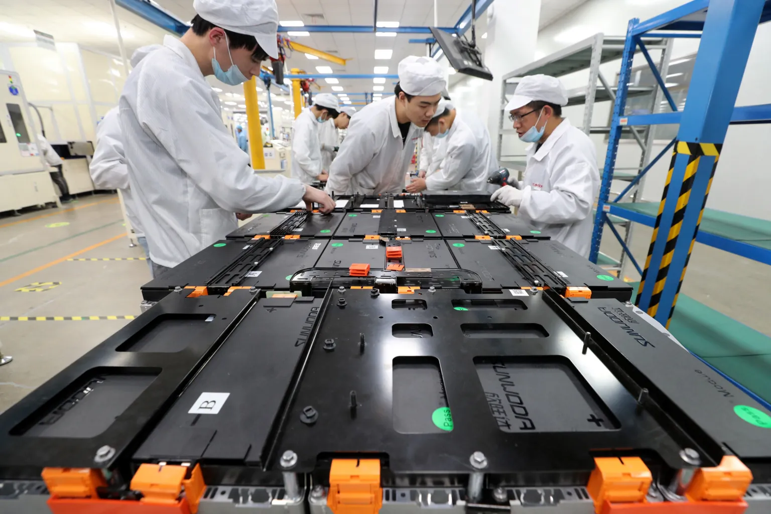AFP via A Chinese factory with batteries open on a work table, workers in white lab coats and wearing blue masks are gathered round the batteries. The frames of the building are painted yellow and blue, with yellow and black warning tape on them. 