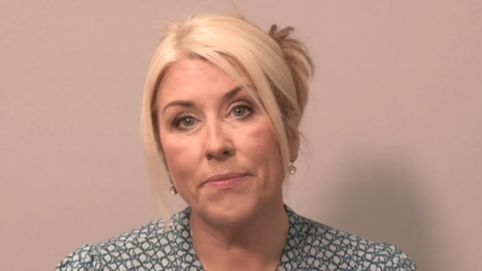 Shows a woman with blond hair tied up in a bun. she is wearing earrings, a green, black and white top. 