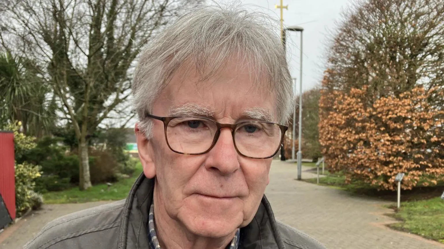 Shows a man with glasses and grey hair and wearing a brown leather jacket standing in front of trees