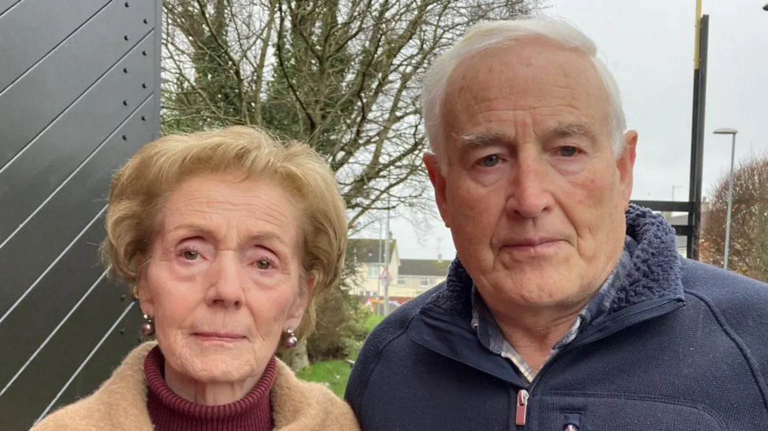 Shows an elderly woman to the left with fair hair, burgundy roll-neck jumper and camel coloured coat. The man to the right is-grey-haired and is wearing a blue zip-up fleece. 