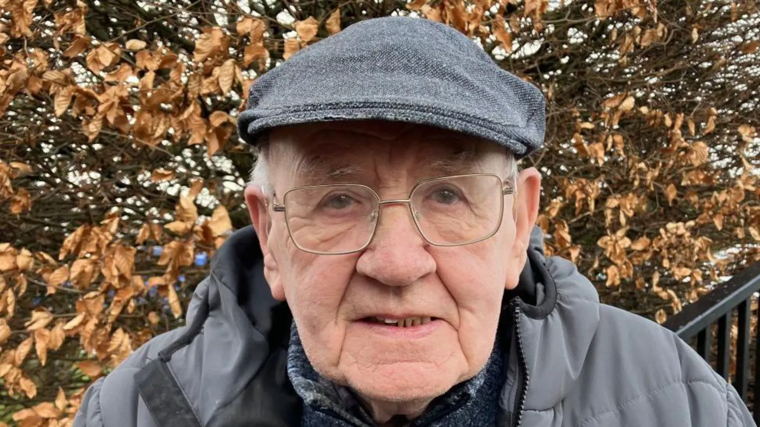 Shows an elderly man with a grey cap, grey coat and glasses