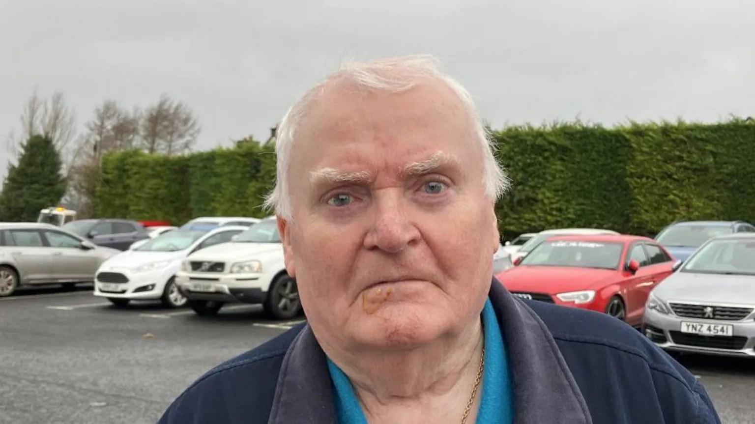 Shows a man with grey hair standing in a car park with cars and a hedge in the background