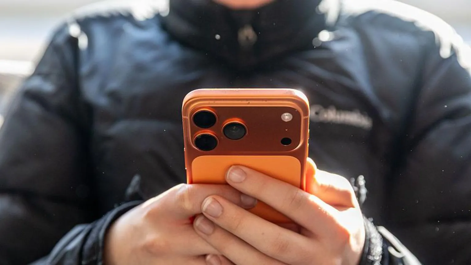 Unrecognisable young boy wearing a black puffer jacket holds a smartphone in front of him.