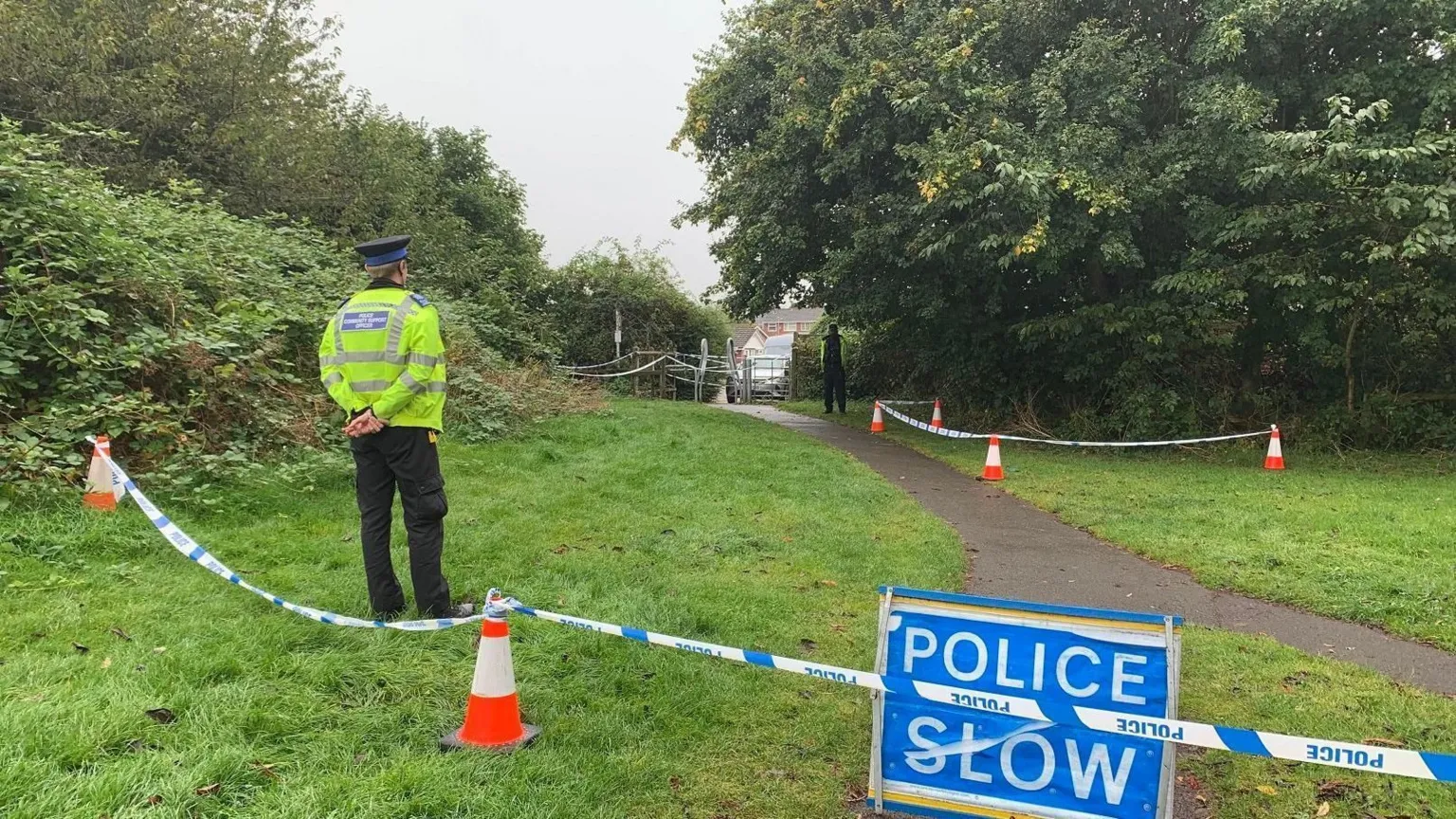 A police officer wearing a hi-vis jacket and hat stands on grass within a cordon in Franklin Park. 