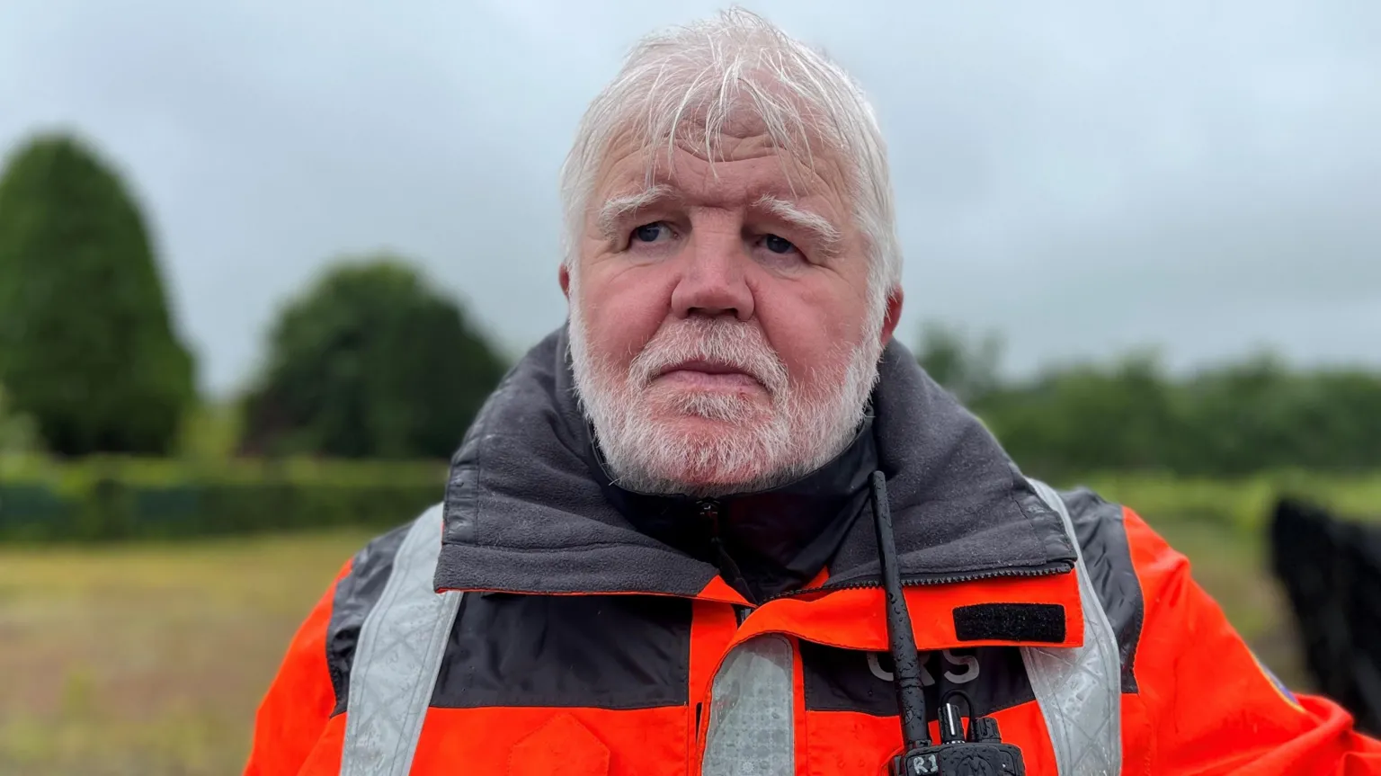 Sean looking at the camera. He is wearing a high vis orange jacket. He has grey hair and a beard. He is standing outside and there are trees in the background.