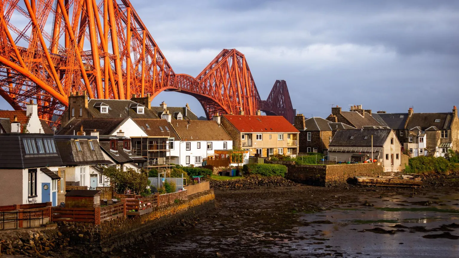 The Forth Rail Bridge stretching behind a row of white and brown houses at sunset.