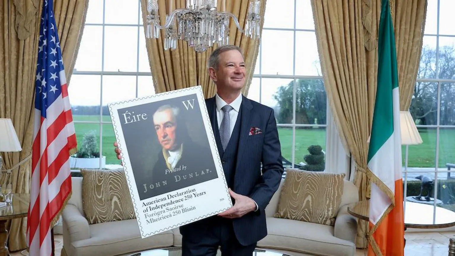 An Post Ed Walsh is holding a large portrait of the stamp. The US and Irish tricolour flags are on each side of him. A sofa, table and lamp are in the background. A lawn can be seen through windows. 
