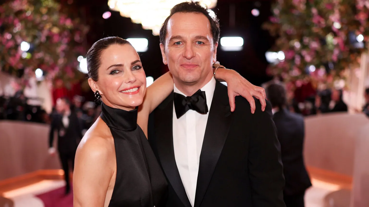  Russell and Rhys on a red carpet, she has her arm around his shoulders. Russell is wearing a black dress with her dark hair slicked back and Rhys is in a suit, with white shirt and a black bow tie. 