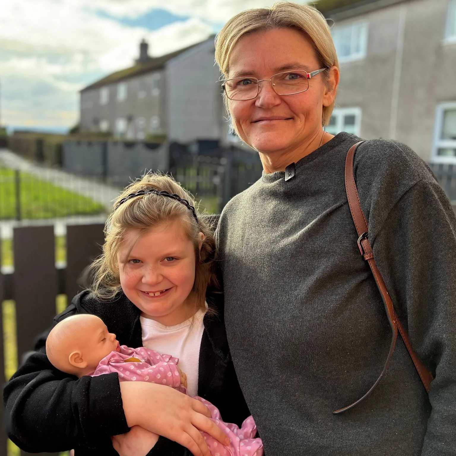 A young girl holding a doll, standing next to a woman with blonde hair and glasses