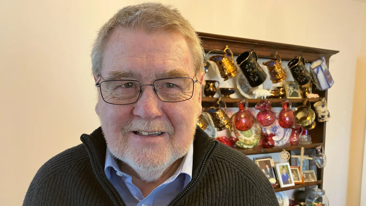 Edwin Roderick, who has short grey hair and a short grey beard, wears a blue shirt and dark jumper. He also wears rectangular glasses. A wooden kitchen dresser can be seen behind him with ornamental jugs and plates on. 