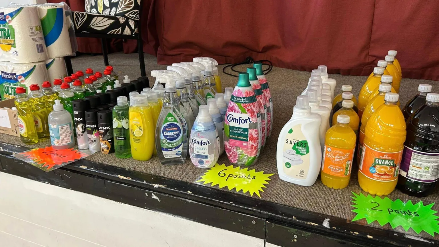 Daventry Community Larder A large number of items on a table including toiletries, drink, all in bottles, all on a stage floor with points written in front. A red cloth is behind the goods. 