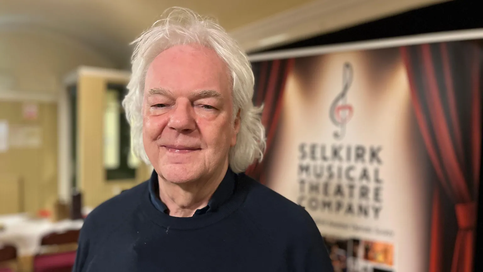A man with grey, shoulder-length hair, wearing a black top, looks at the camera.