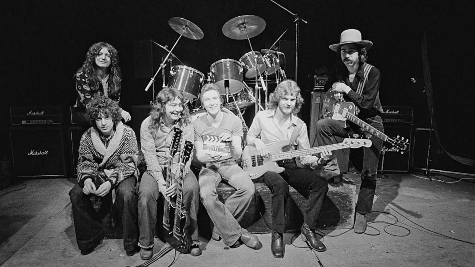 Fin Costello/Redferns/ A black and white photograph of six men, mainly holding musical instruments, sitting in front of a drum kit.