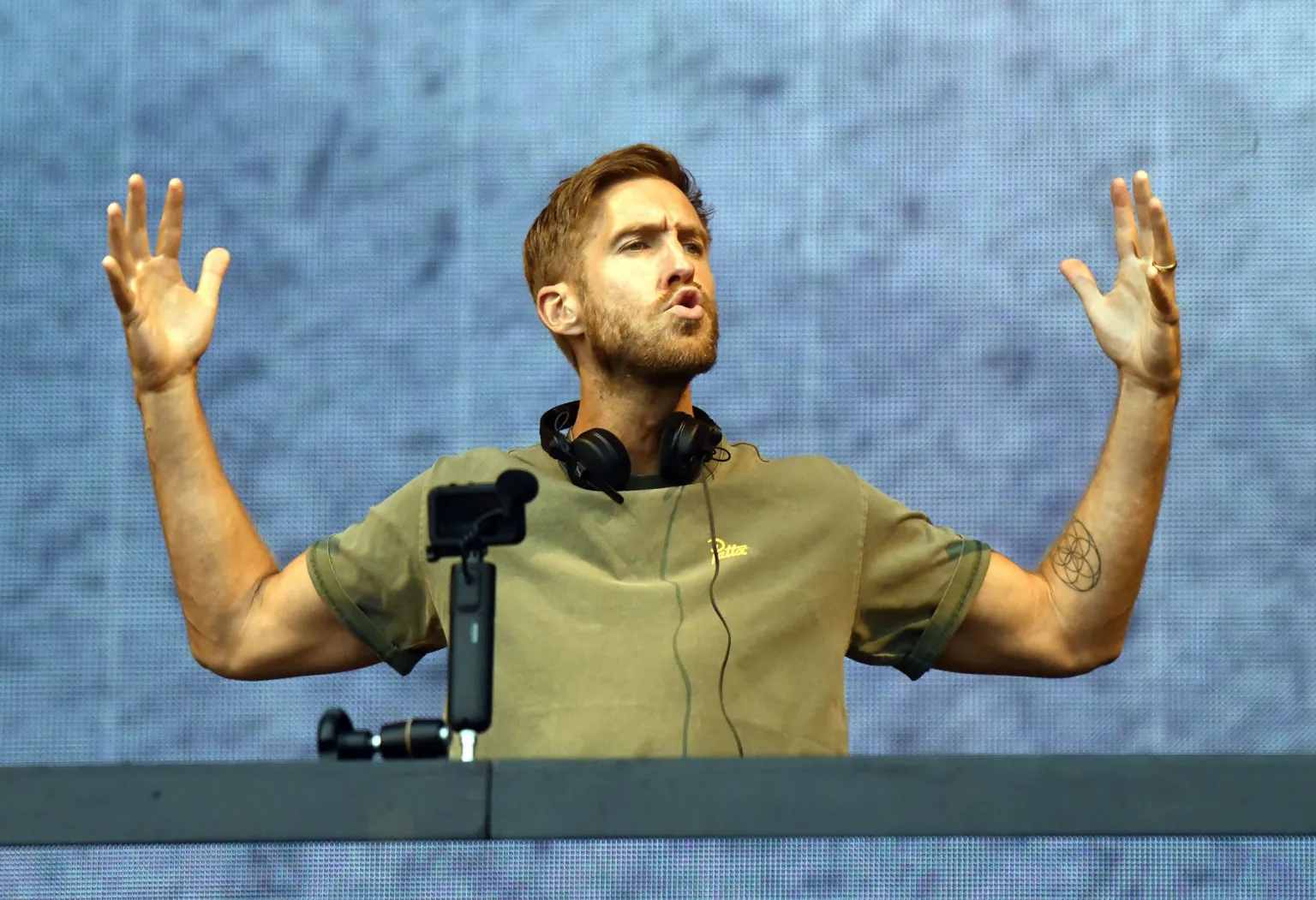  A man in a green T-shirt with black headphones around his neck. He is standing behind a desk and has his arms in the air. 