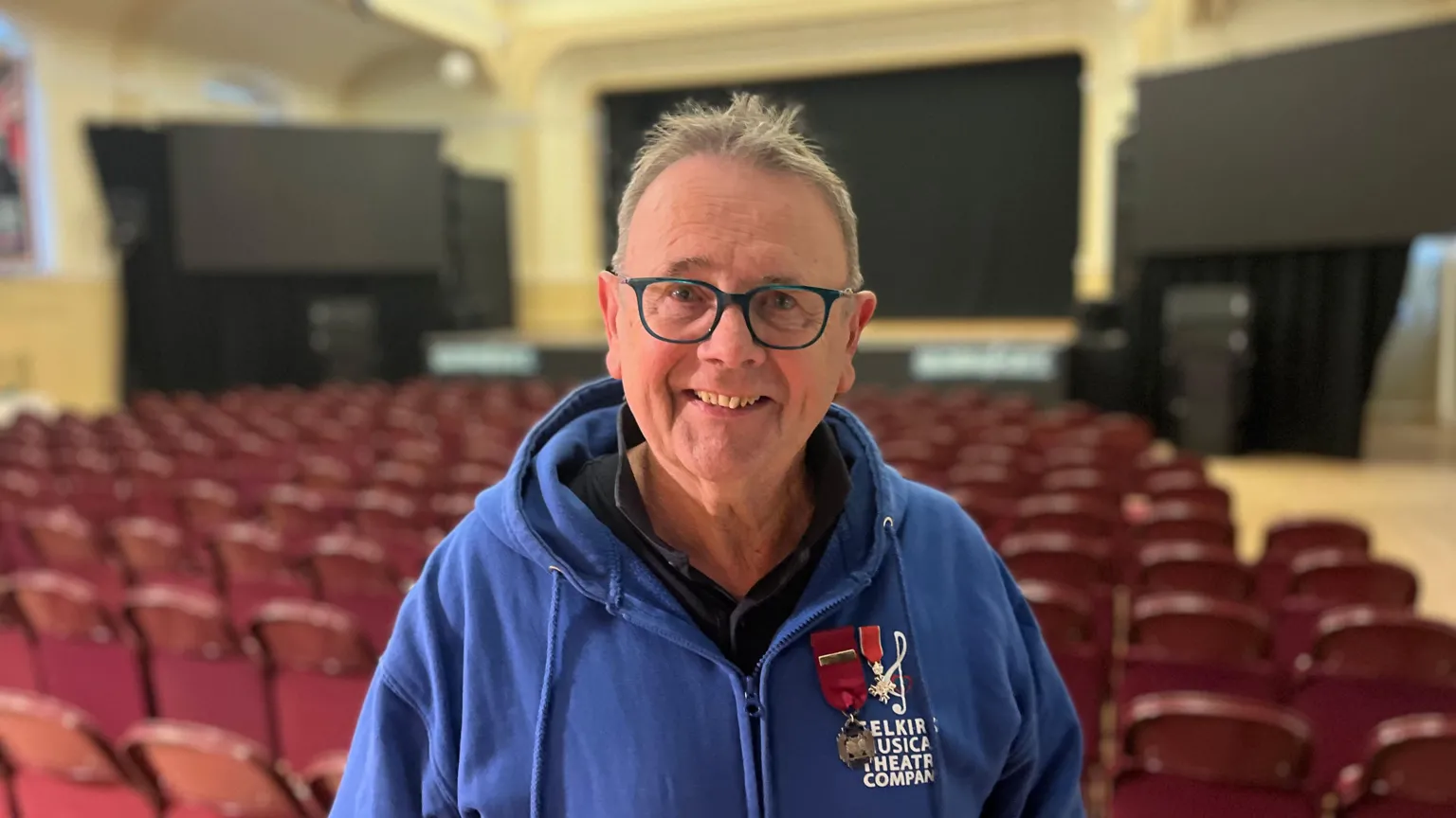 A man with glasses and short hair, wearing a blue hoodie, smiles at the camera. Behind him is an empty theatre