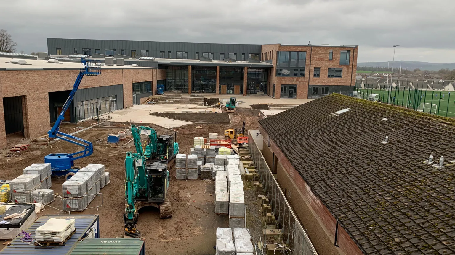 A new school building viewed across a bit of a building site with construction materials and vehicles on muddy ground