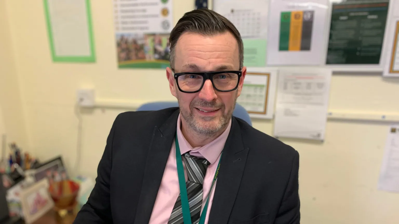 A man with dark hair slicked back with glasses and a slight growth of a beard. He is wearing a pink shirt, a striped tie and a black suit jacket