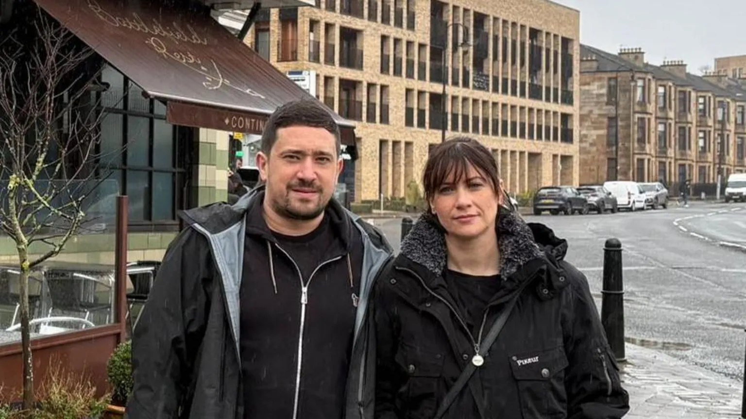 Alex Matheson A man and a woman stand outside a restaurant on a busy street. It has been raining and the road is wet, both are looking directly into the camera and have dark hair.