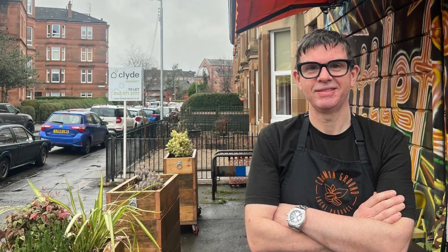 Neil Bolland A man with dark glasses stands outside a coffee shop smiling.