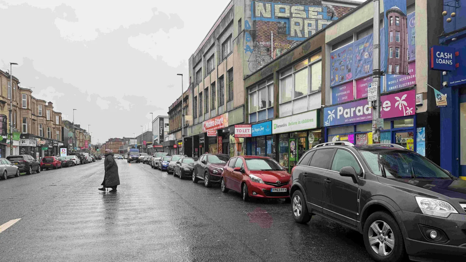 A busy main road with shops and parked cars. A woman is crossing the road.