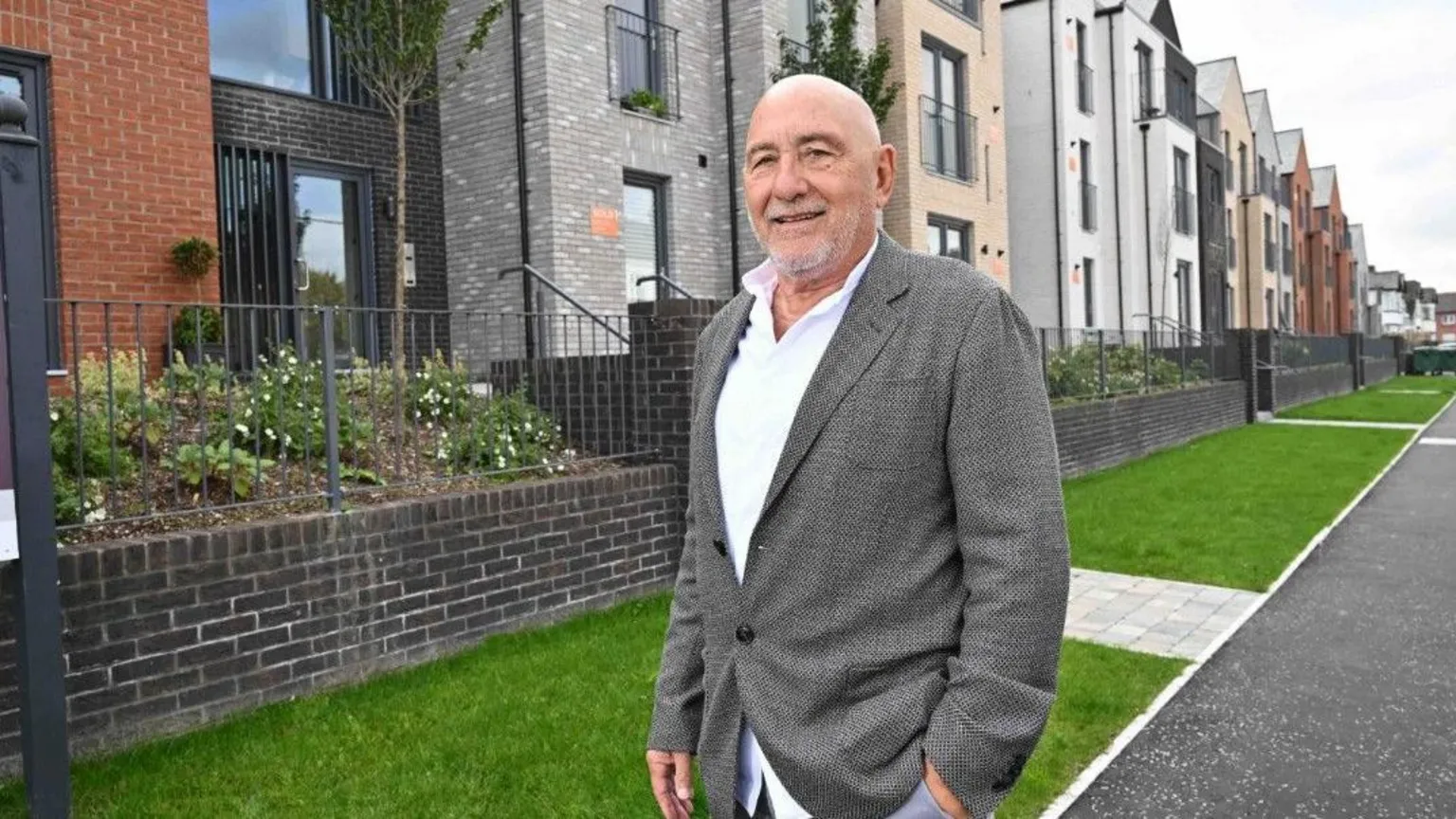 Hagan Homes James Hagan, a man with grey hair and a grey beard, wearing a grey jacket and a white shirt. He is standing in front of various houses and green grass. 