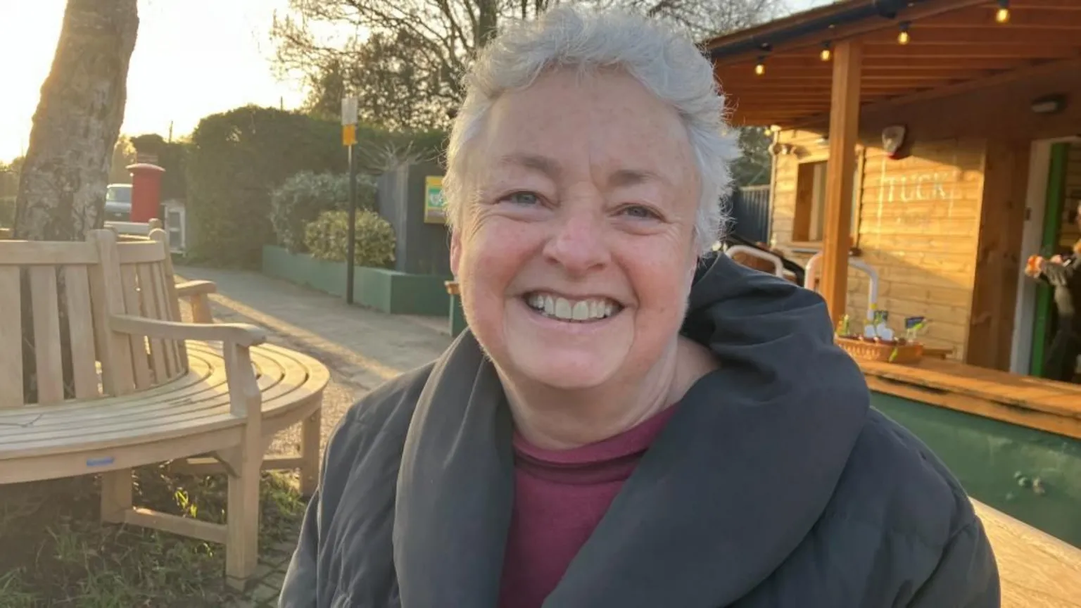 Vikki Irwin/BBC Clare Thomas is smiling. She is standing outside a cafe with a wooden bench to her right that goes around a tree. She is wearing a grey coat and a maroon top underneath.