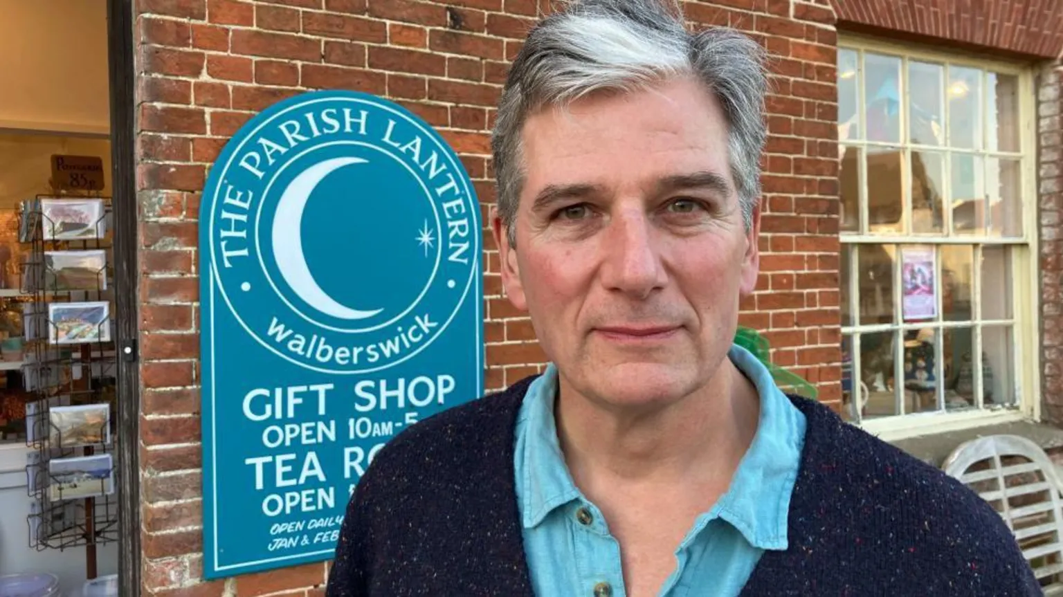Vikki Irwin/BBC Robin McLean stands in front of his blue shop sign for The Parish Lantern gift shop and tearoom. He has grey hair, a dark blue cardigan and a light blue open-necked shirt.