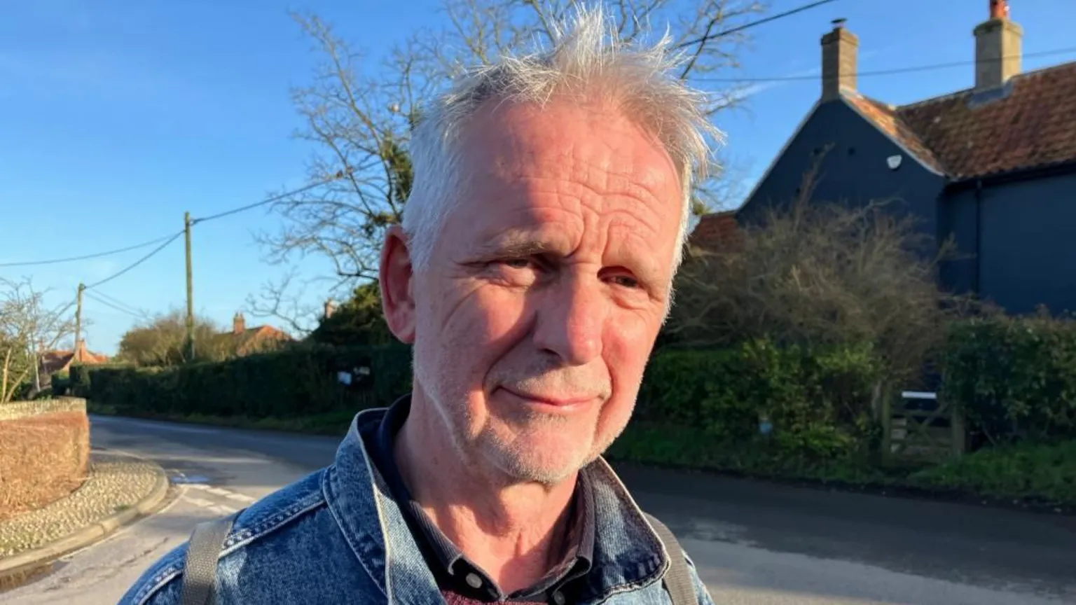Vikki Irwin/BBC Man with grey hair stands in the village on a street corner with a brick wall to the side and a black coloured house behind. He wears a denim jacket and the sun is shining on his face. There is a blue sky and telegraph poles behind him