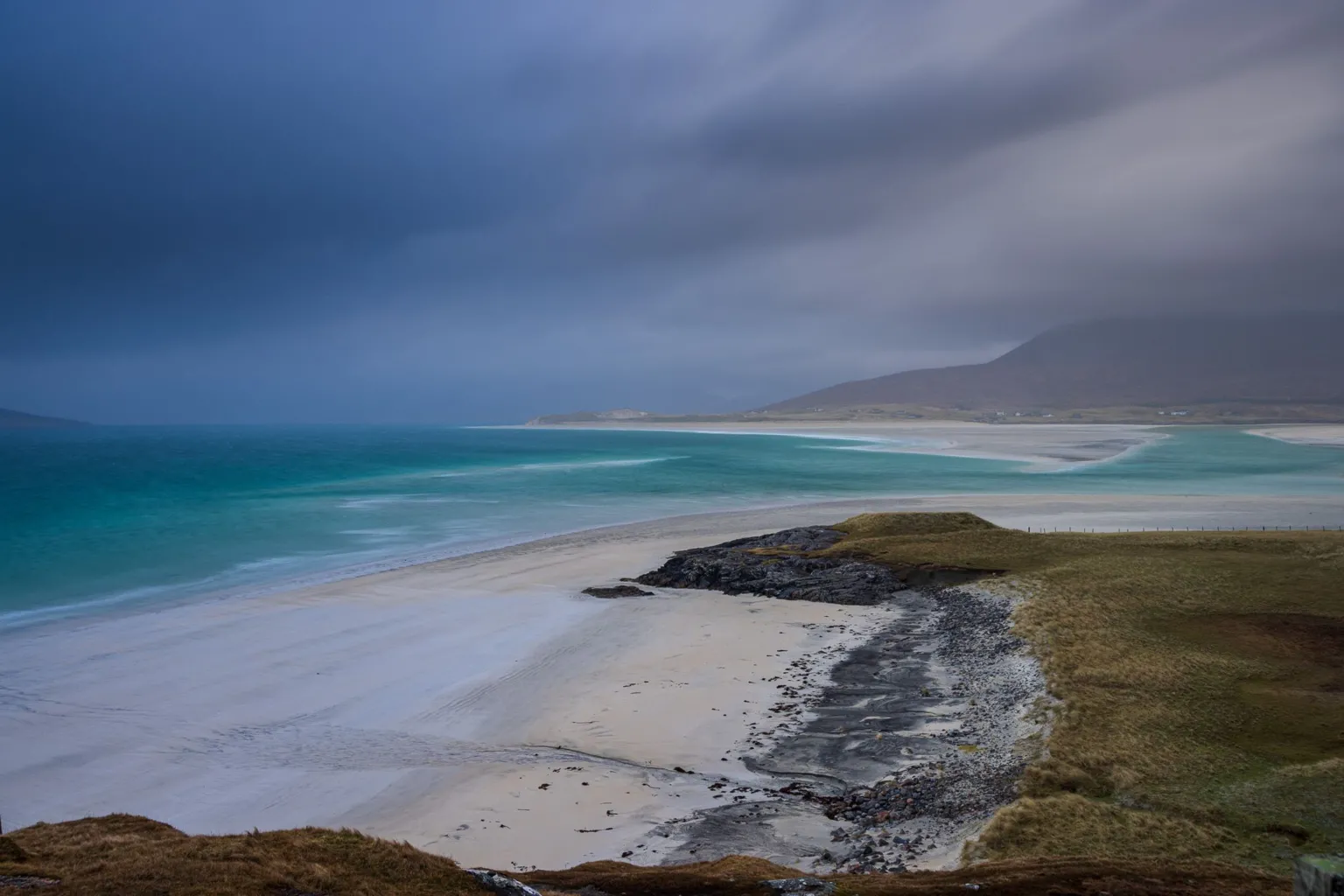 Andrew Briggs A beach on the Isle of Harris. The water is a turquoise colour and the sky is dark with clouds. There are hills in the background.