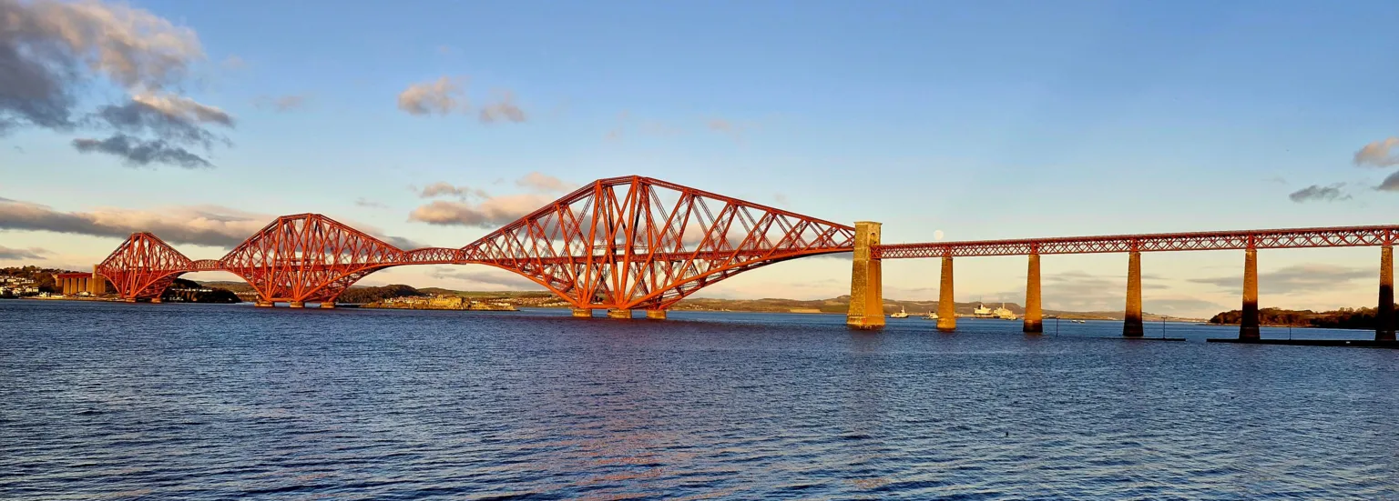 Erin Harper The Forth Rail Bridge over the Firth of Forth. The bridge has three cantilevers and is painted red. The water has ripples in it and there are a few clouds in the sky.