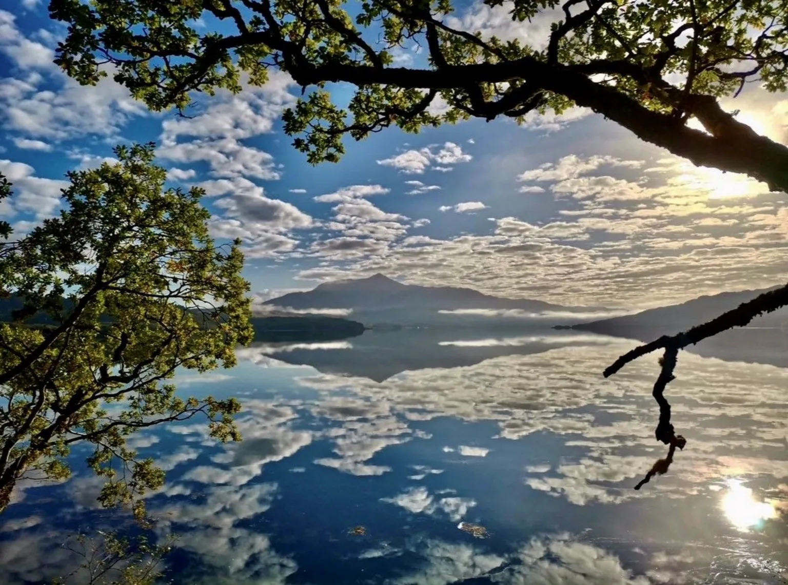 Richard Castro Beinn Resipol reflected in Loch Sunart. The sky is bright with patches of cloud that are reflected into the loch. Trees frame the mountain and the sun sits in the top right corner.