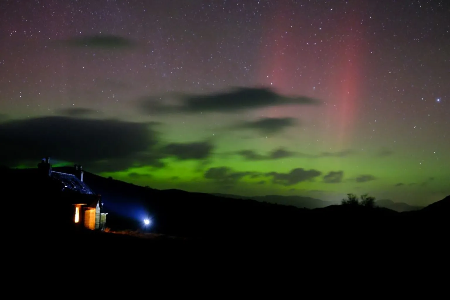 Sam Morris The Northern Lights in the Scottish Highlands. The sky is green with some dark clouds and red stripes. The stars are visible.