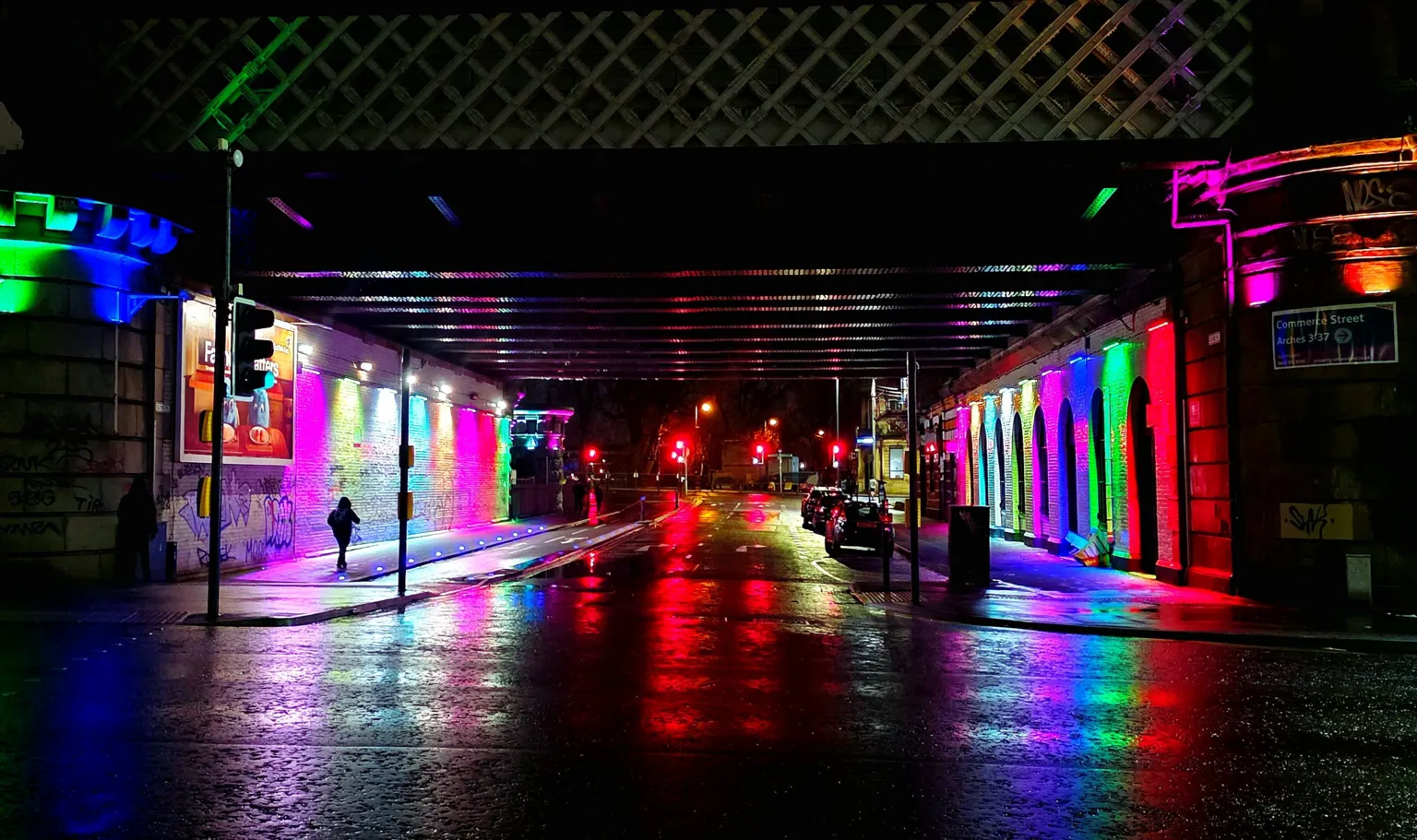 Ruth Cumming A city street on a wet and dark night. Various lights - purple, red, green and blue - are reflecting across both sides of the road. 