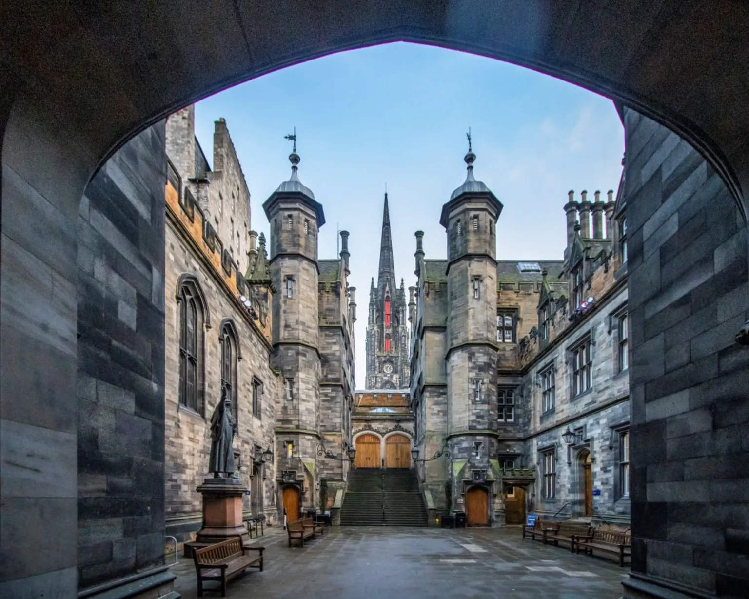 Steven Neish Gothic-style brick buildings with turrets and spires. The image is taken from through a tunnel of an empty quadrangle at the University of Edinburgh. There are benches in the quad and statues around the edges.