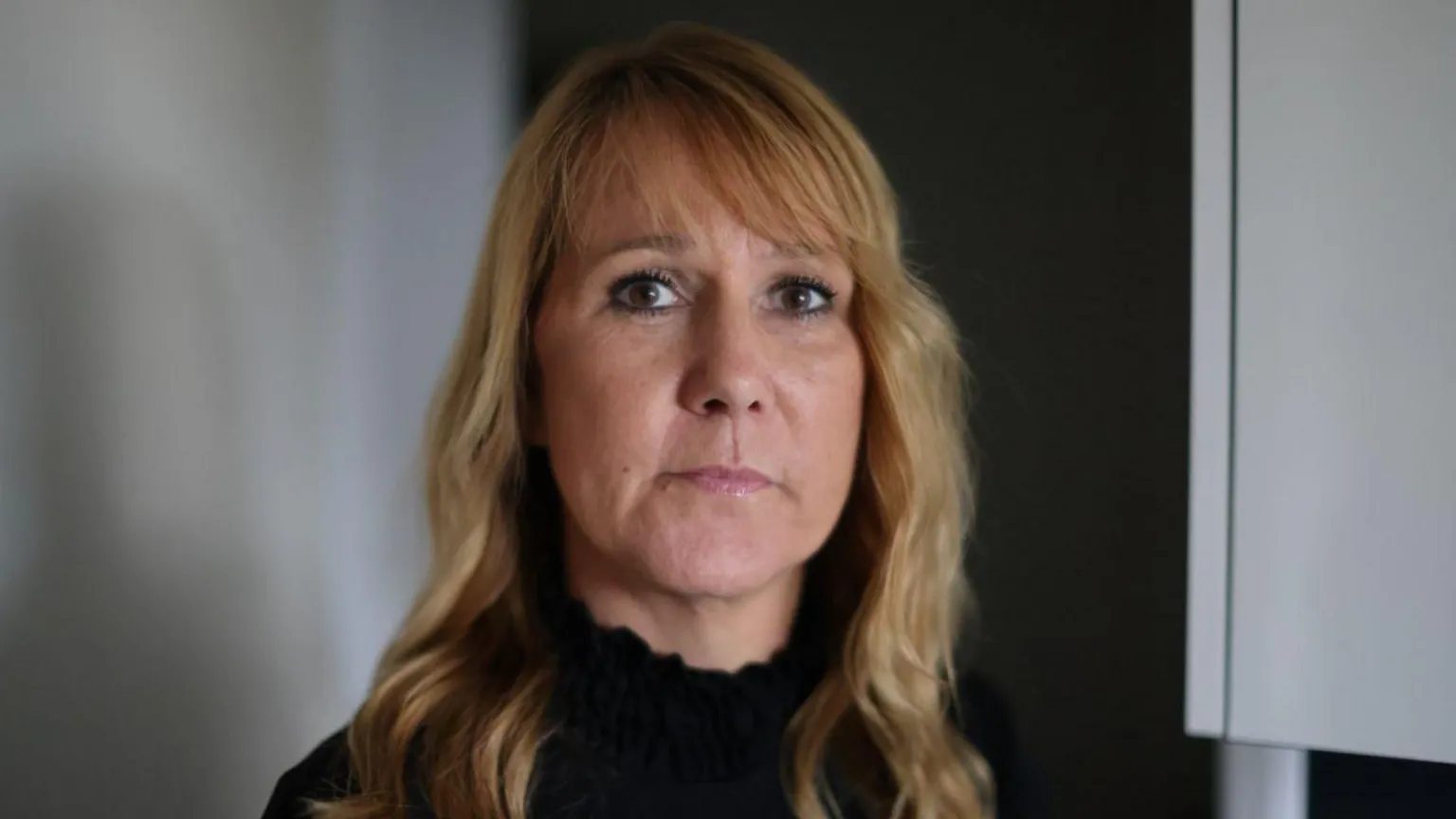 A woman with blonde hair and brown eyes, wearing a black jumper, looks at the camera