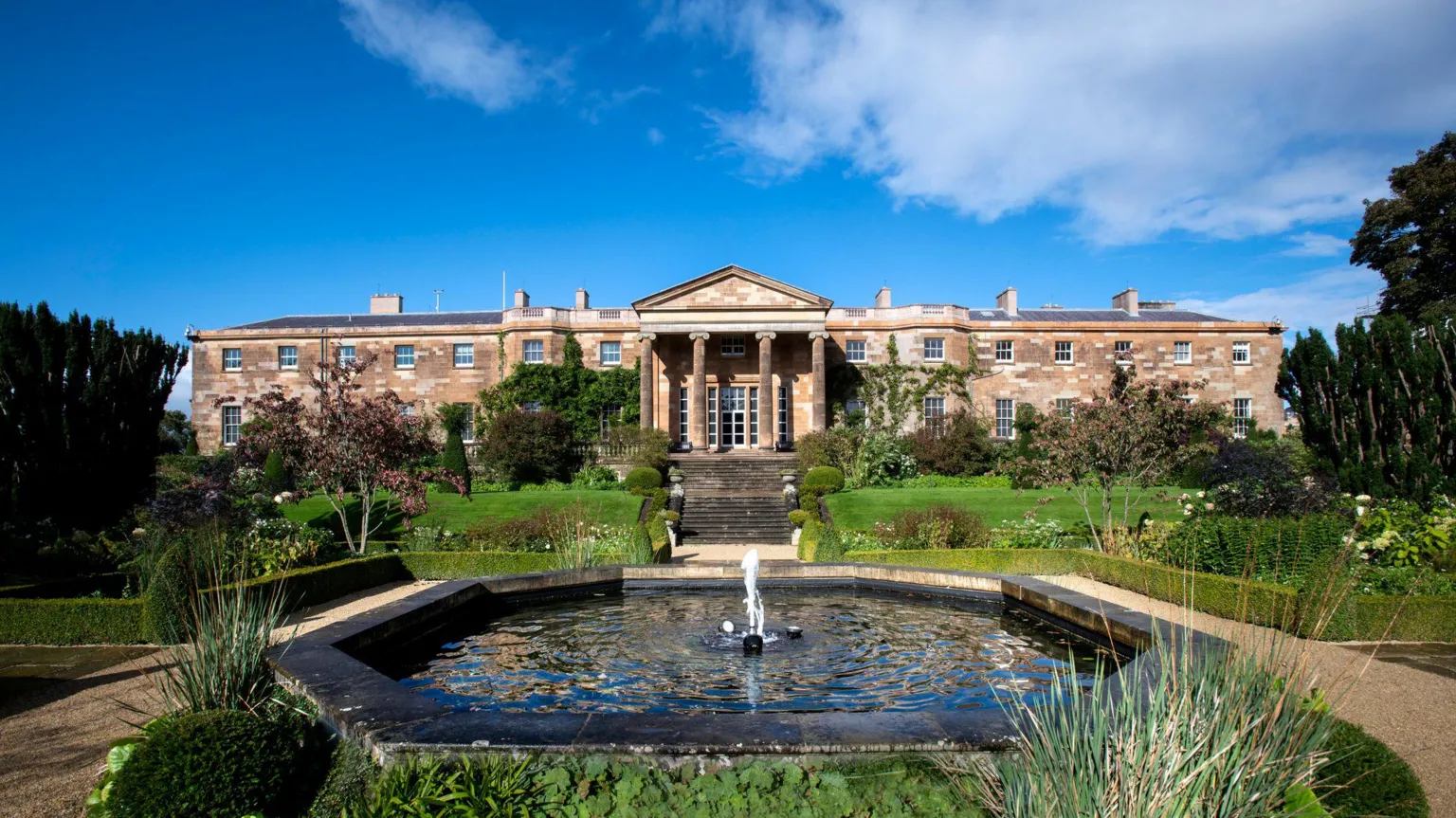  Hillsborough Castle building, with a pond in front of it. There is a blue sky above. 