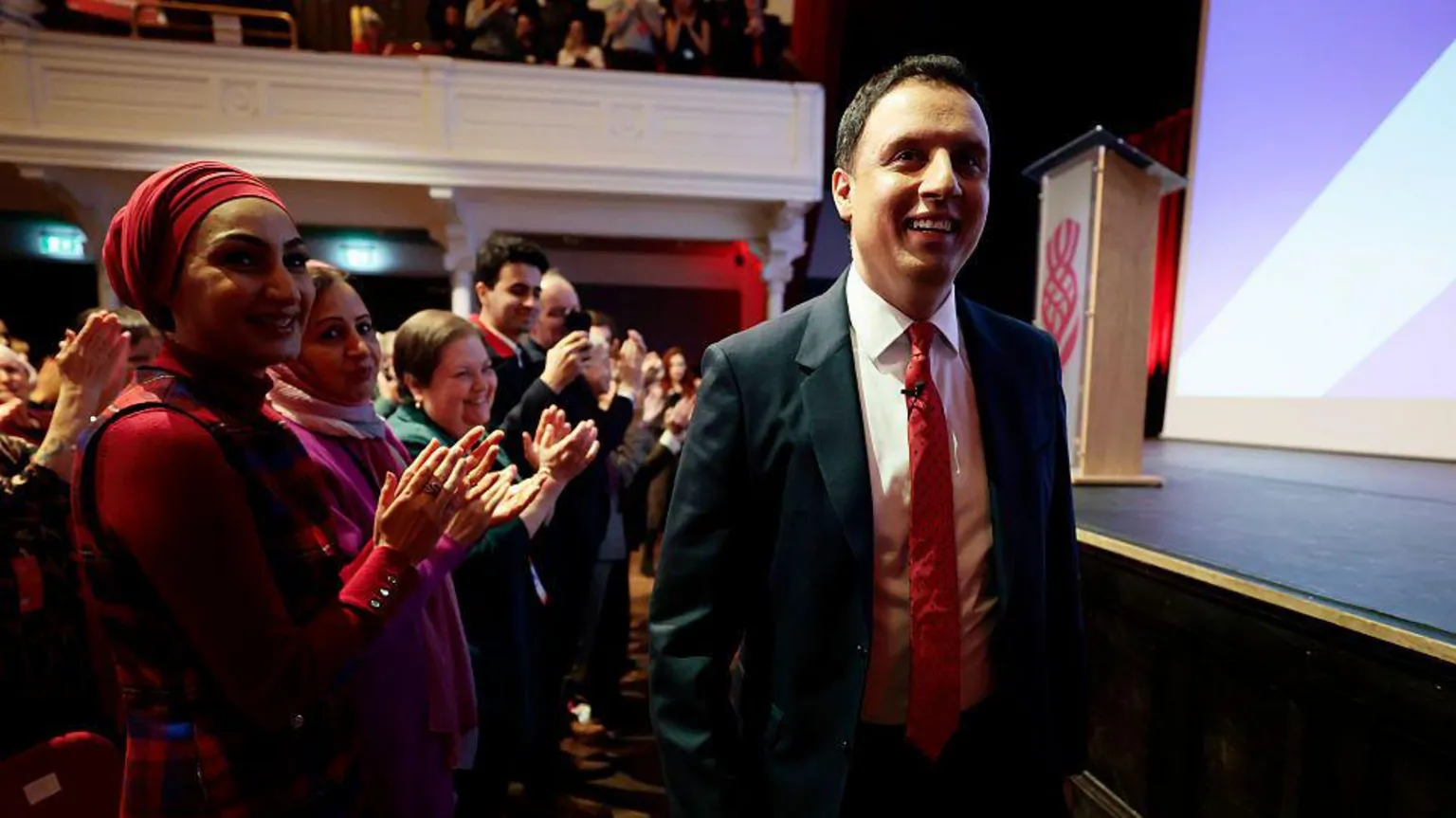  Anas Sarwar wears a dark suit with white shirt and red tie. He has short dark hair and walks by the front row of a theatre by the stage. People stand up clapping him 