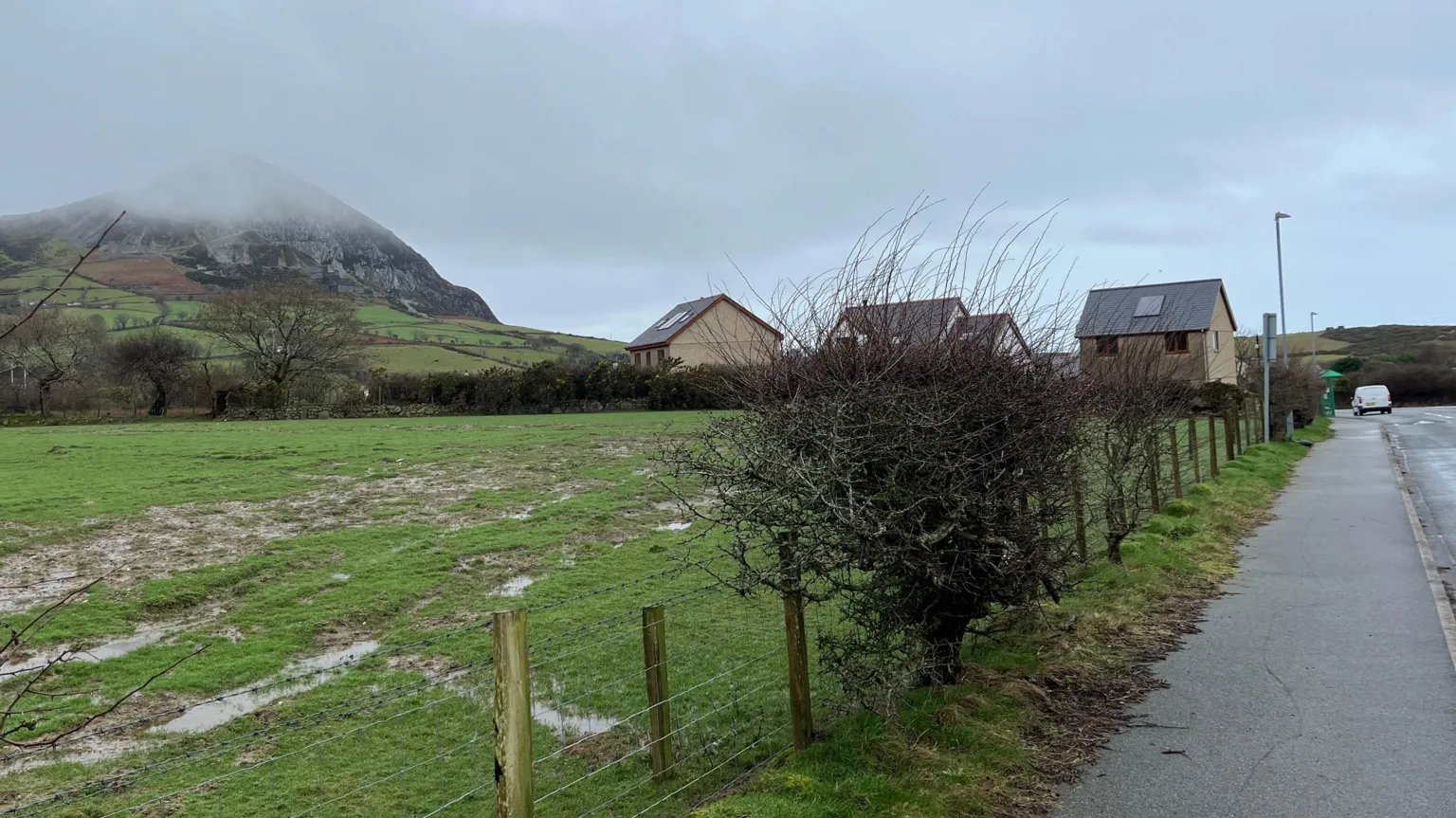 A picture taken from the side of a road where the development is proposed. it is a green field with several houses visible behind a windrow hedge. a mist covered peak is in the distance
