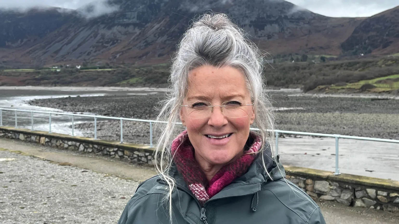 a woman looks into the camera. sh has long grey hair and is wearing a green parka done up over a red scarf and she has rimless glasses. in the background is a stone wall with a green metal railing above a large body of water with mountainous peaks in the distance 