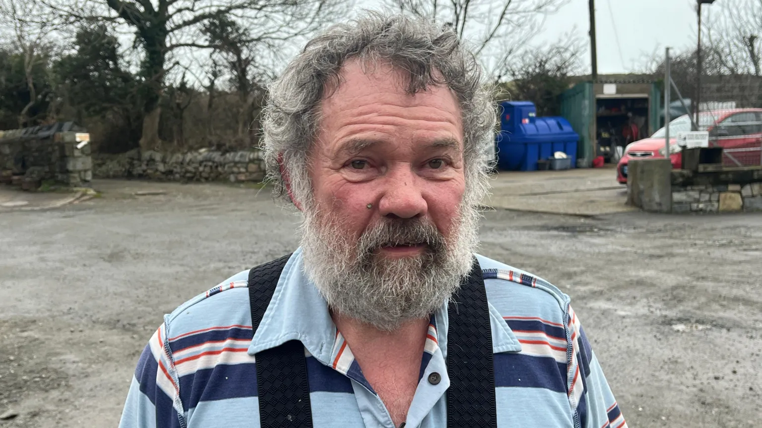 man looking at the camera. h has a grey beard and medium length grey hair. H has on a t shirt with blue and red and white stripes. There is a garage in the background beside a stone wall and trees. 