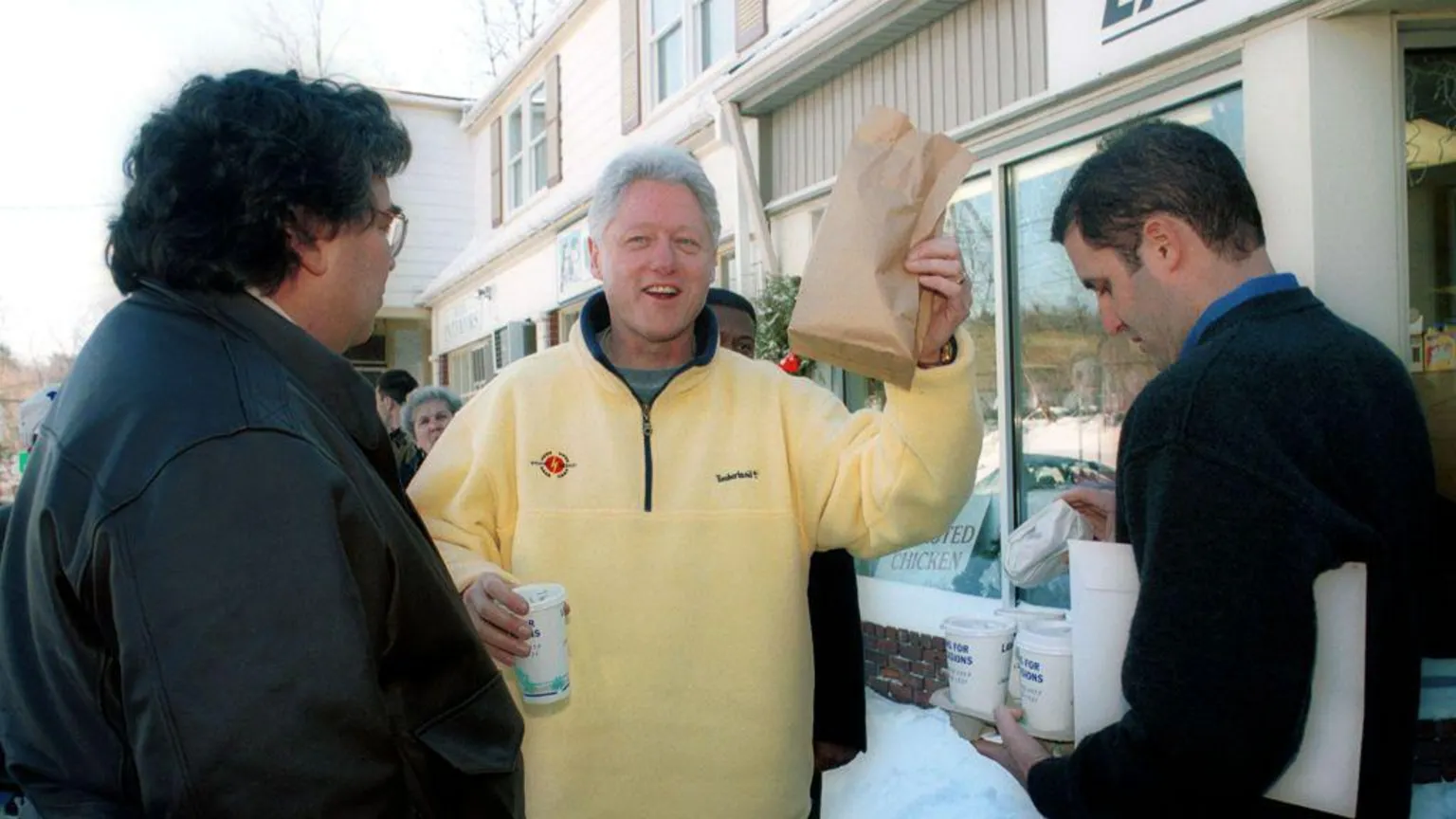  Band, wearing a black jacket, stands on the street holding several takeout coffees while Clinton, wearing a yellow sweatshirt, triumphantly holds up a brown paper takeout bag and carrying a to-go coffee